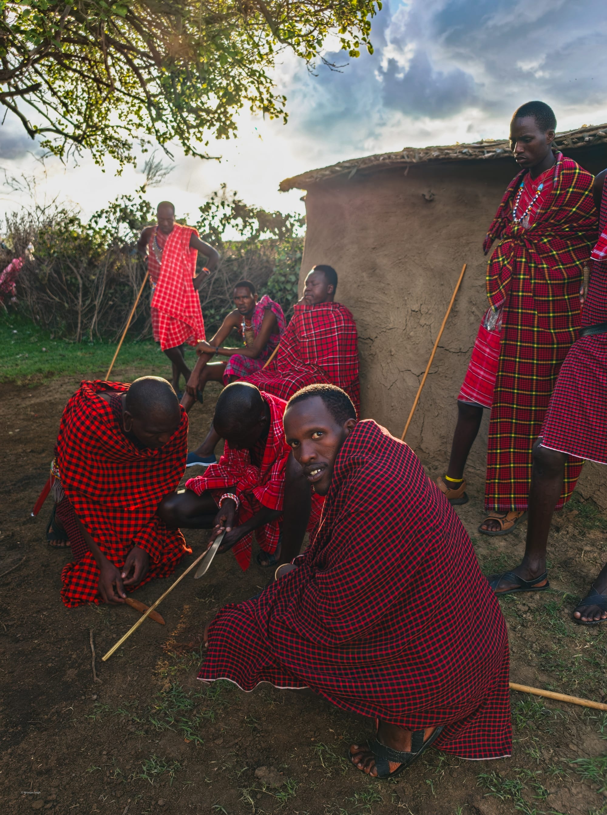 Maasai men in red shukas crouched on the ground starting a fire with tinder wood outside a mud-walled house near Sekenani