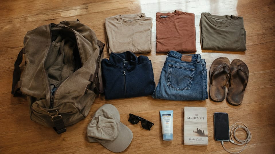 Flat lay of minimal safari packing on a wooden floor: a khaki duffel, folded t-shirts, a fleece, jeans, leather flip-flops, cap, sunglasses, sunscreen, a book, and a phone.